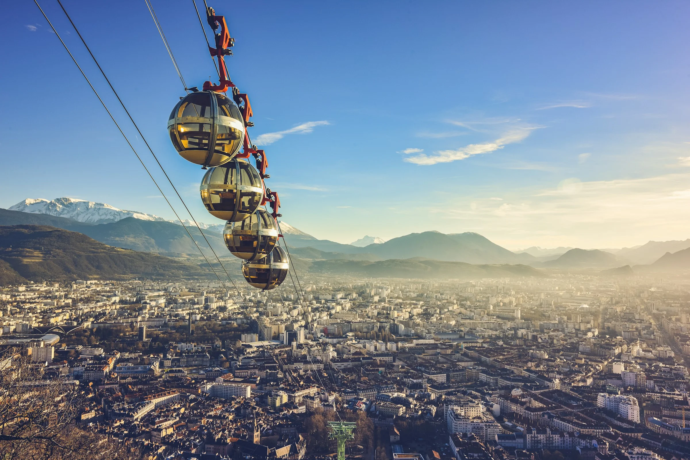 Aerial view of Grenoble, France, featuring the iconic bubble cable car against the backdrop of a city sprawl, distant mountains and a clear sky
